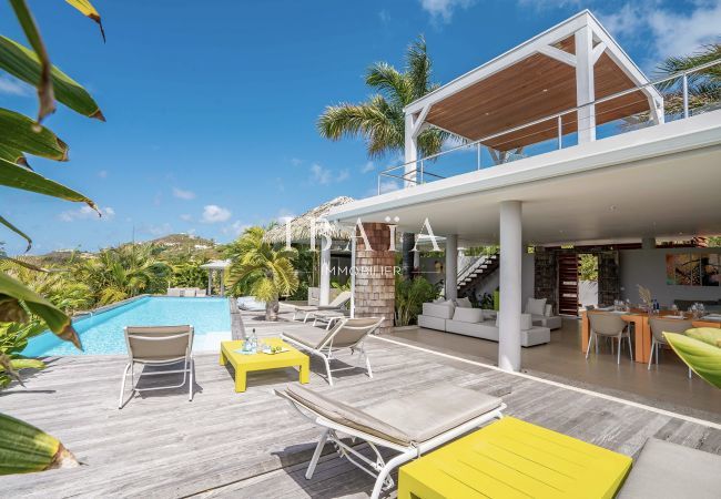 Vista de la terraza de madera con 4 tumbonas, mesas de café, piscina y terraza sombreada con salón exterior y mesa de comedor