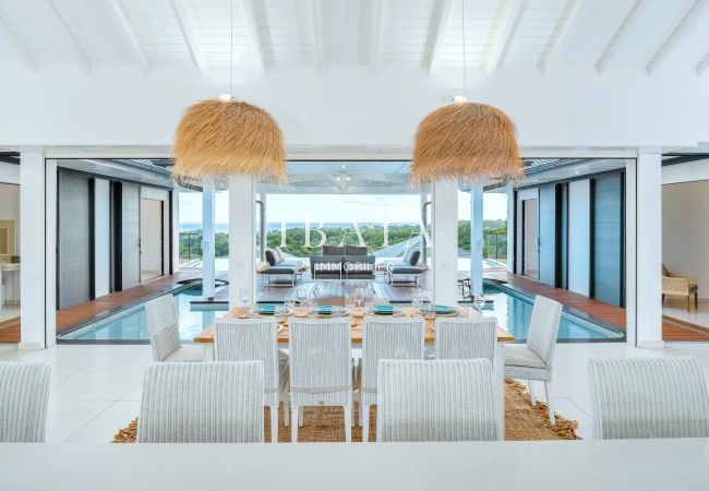 Espacio de comedor al aire libre con sillas blancas, lámpara de paja y vista a una piscina y sofás.