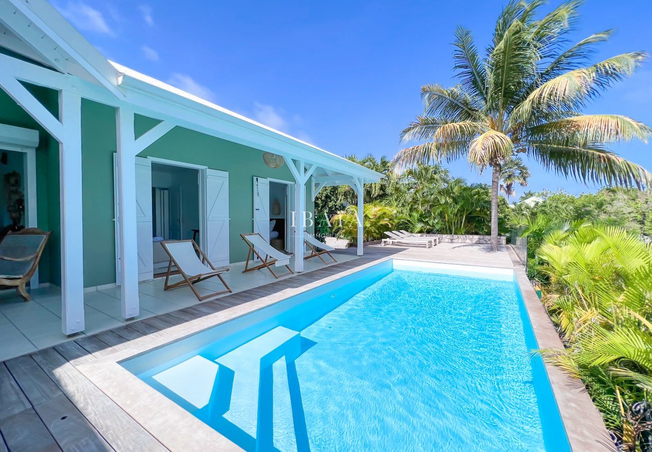 Vista de la piscina y la terraza de madera con las habitaciones con las ventanas francesas abiertas, en una villa de alta gama en las Antillas.