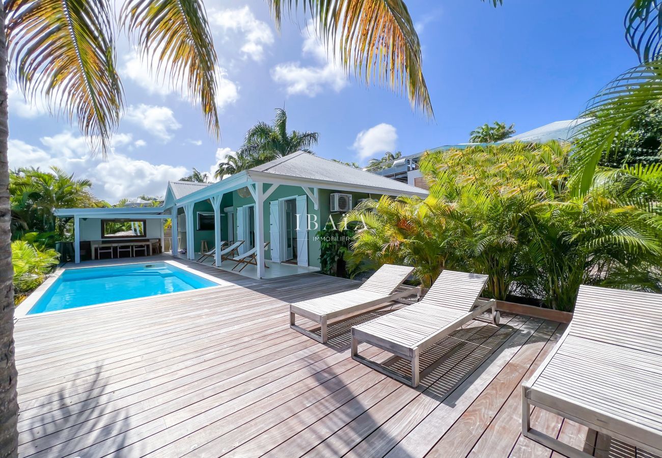 Vista de la terraza de madera, la casa, las tumbonas de madera y el comedor de madera en un jardín tropical, en una villa de alta gama en las Antillas