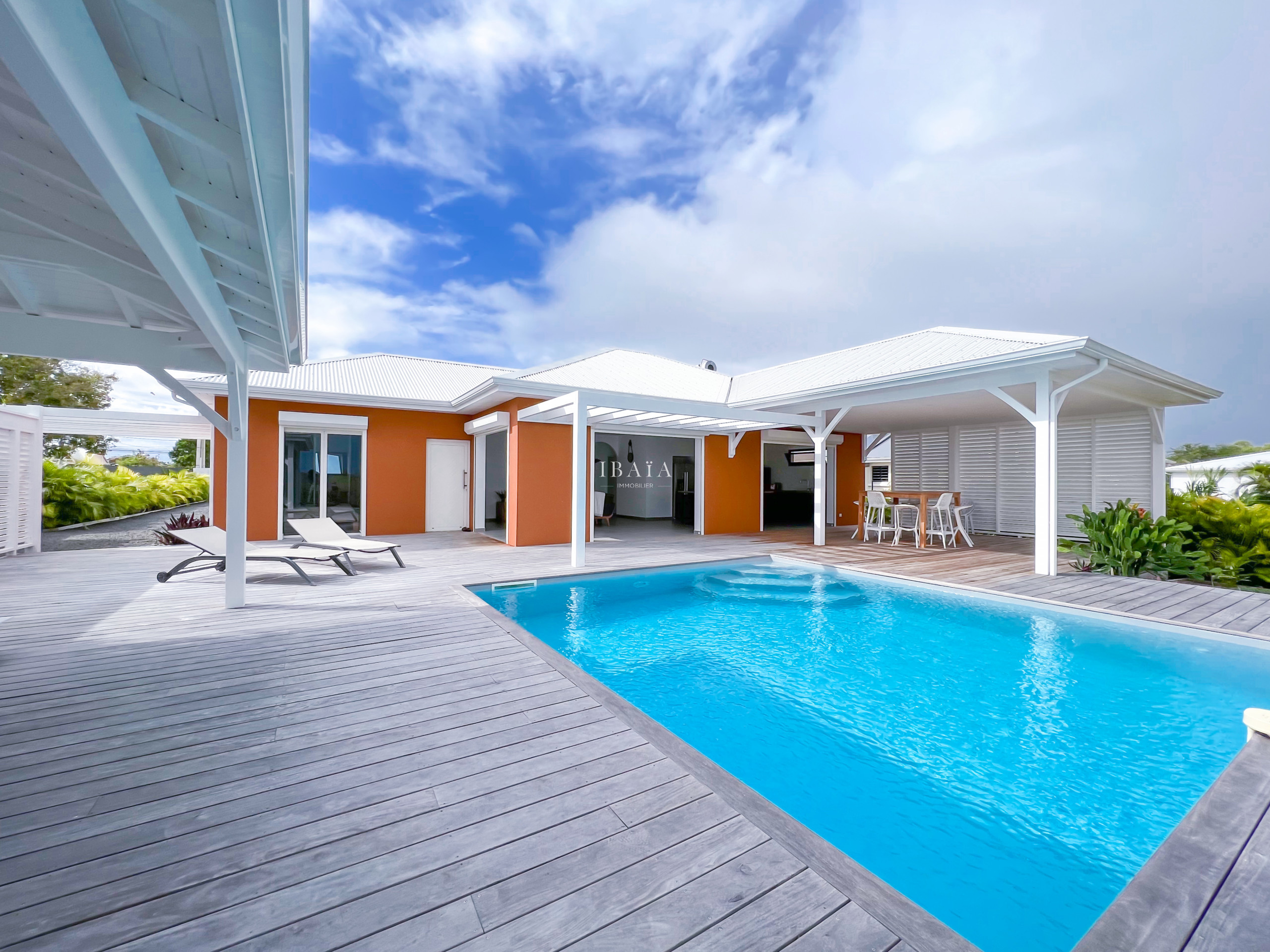 Vue de la terrasse en bois avec piscine, transats, table haute et chaise haute dans une villa haut de gamme aux Antilles