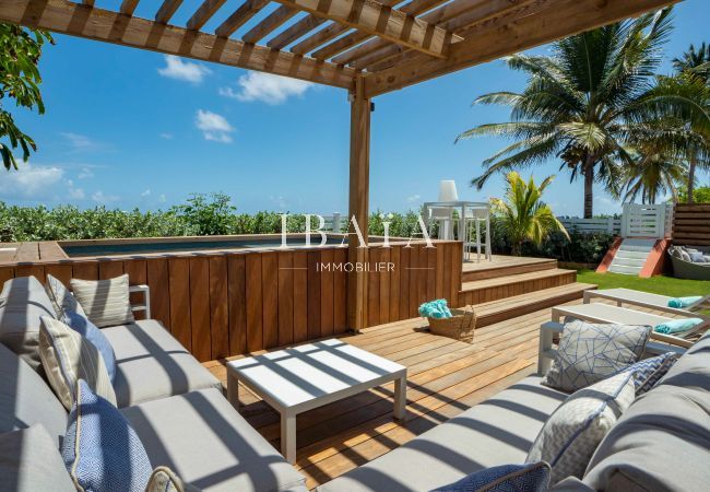 Vue du salon de jardin à côté de la piscine surélevée avec coffrage en bois, sous un patio ombragé en bord de mer