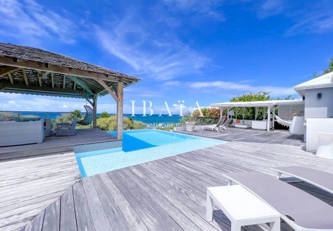 Vue de la terrasse avec patio ombragé et salon de jardin, piscine, terrasse en bois et transat offrant une vue sur la mer dans notre villa haut de gam