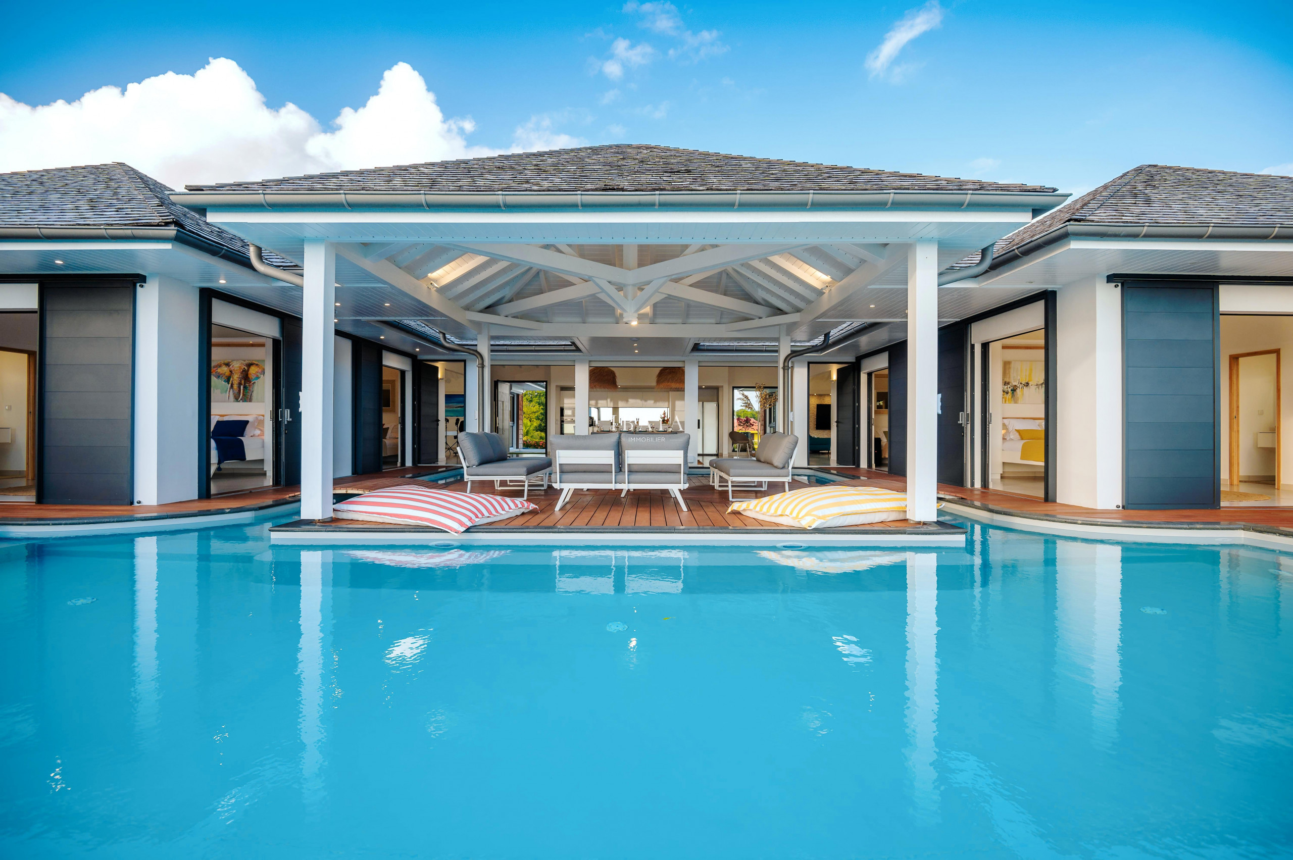 Maison de luxe avec piscine turquoise, terrasse en bois et intérieurs visibles.