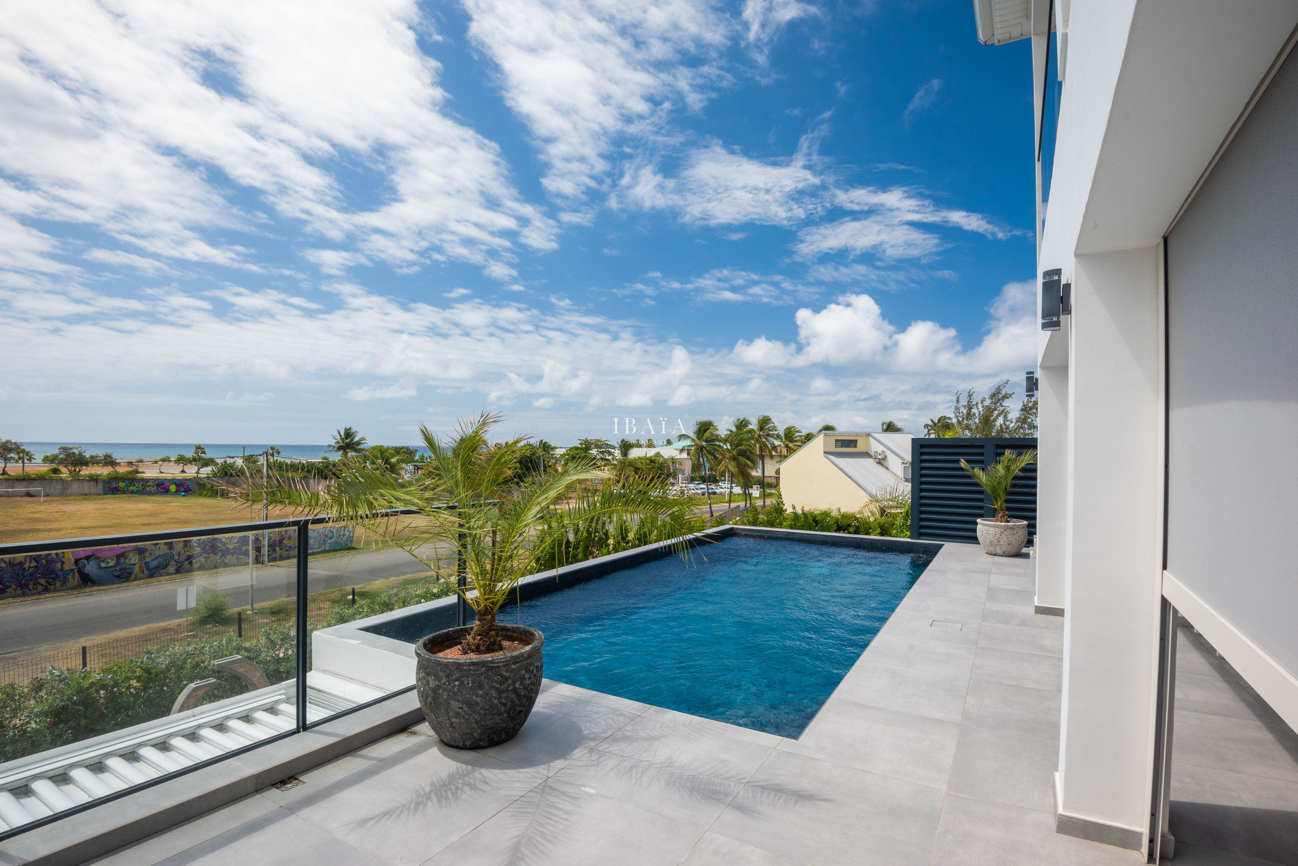Vue d'une terrasse avec piscine donnant sur la plage et le ciel bleu nuageux.