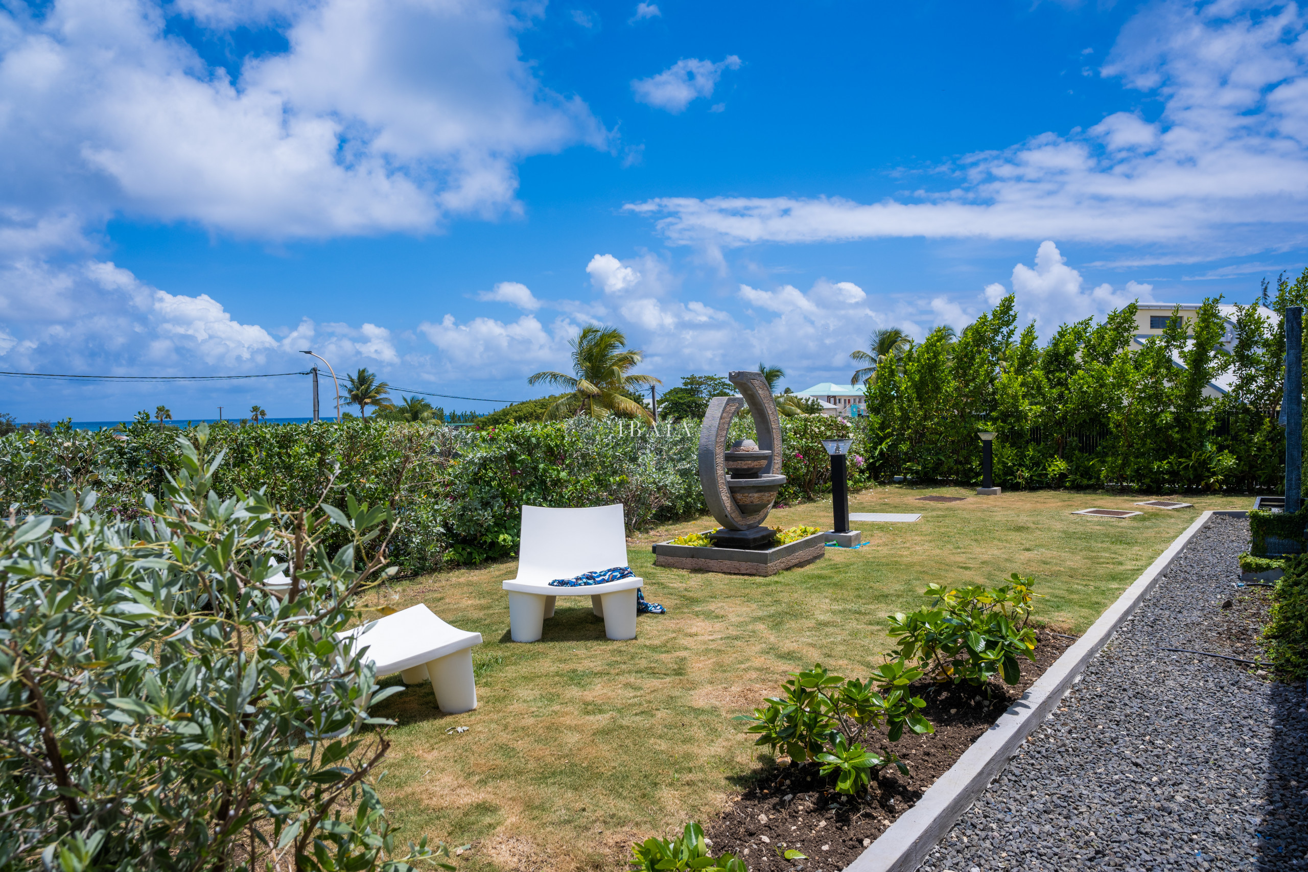 Jardin tropical avec une sculpture moderne, une chaise blanche et vue sur l'océan.