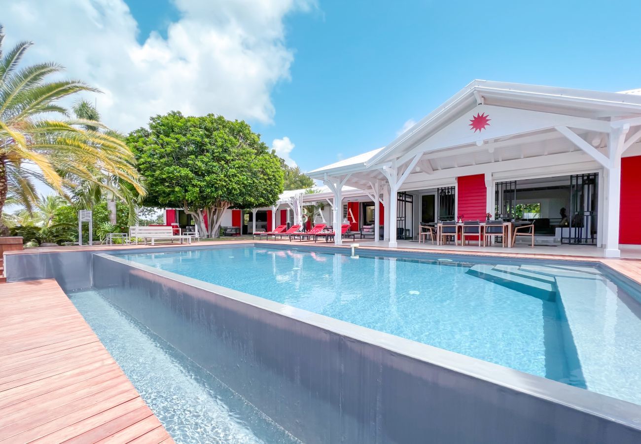 Vue sur la piscine à débordement, la terrasse spacieuse et la façade de la maison avec table d'extérieur et transats rouges dans une villa haut de gam